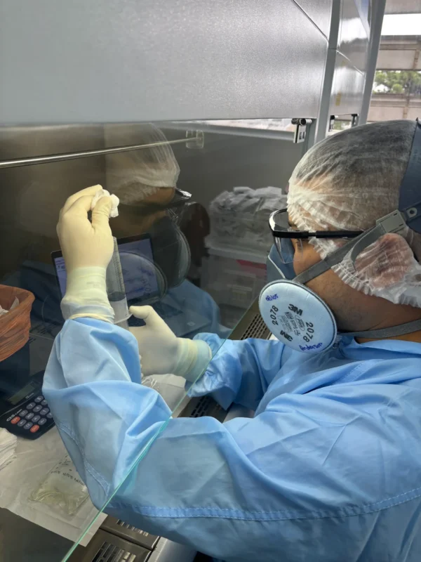Profissional do IOP utilizando cabine de segurança biológica para manipulação de amostras laboratoriais, com uso completo de EPI e соблюindo normas de biossegurança.